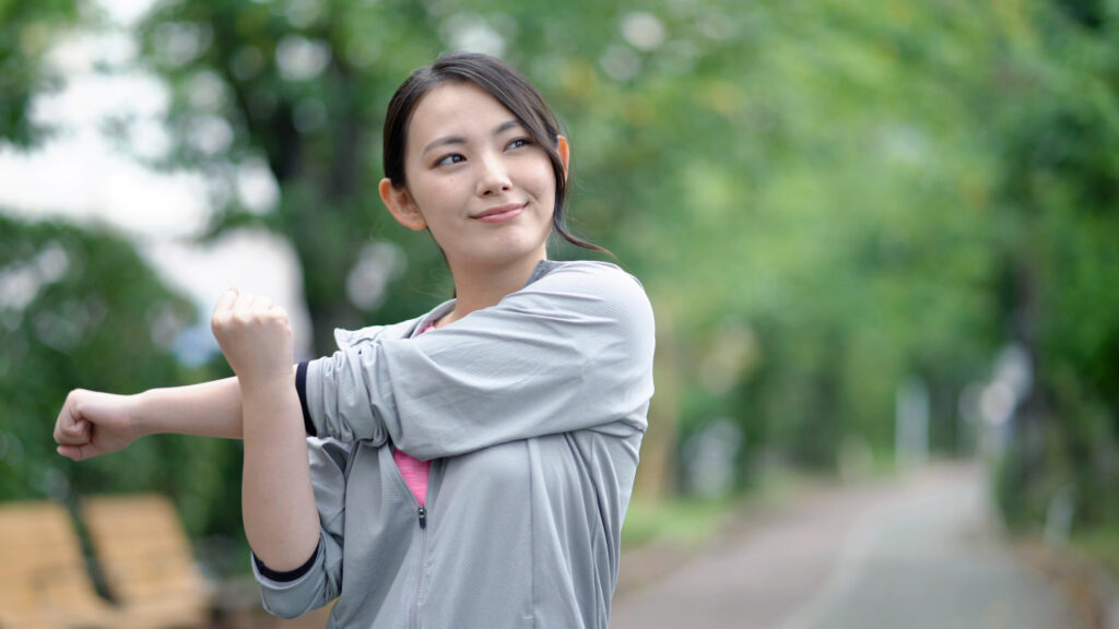 朝の公園でストレッチをする若い女性。肩と腕を伸ばし、気持ちよさそうにウォームアップしている様子。鹿児島荒田のまどか整体院