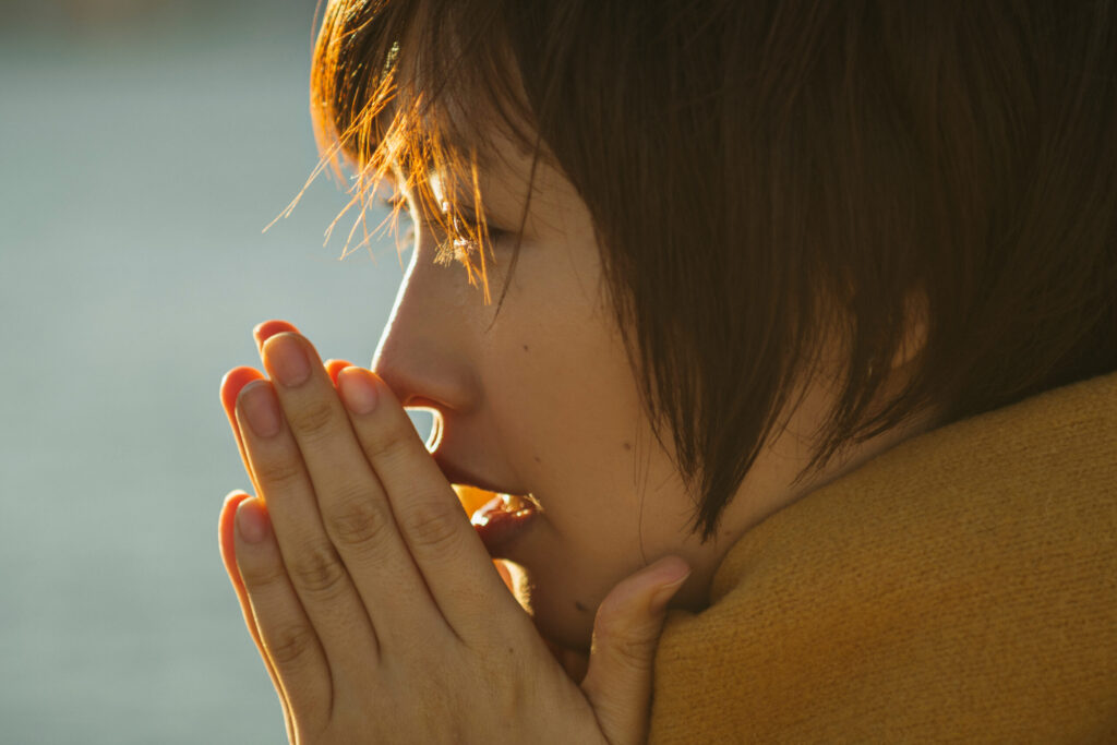 寒さの中で手を合わせて息を吹きかける女性の横顔。冬の冷え込みを感じる様子(鹿児島荒田のまどか整体院)