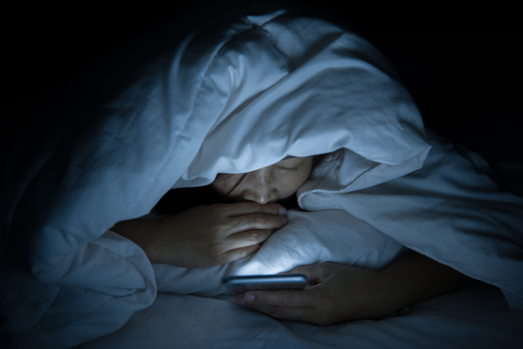 布団の中でスマートフォンを見続ける女性。夜のスマホ習慣が自律神経を乱し、更年期やPMSの不調につながることも