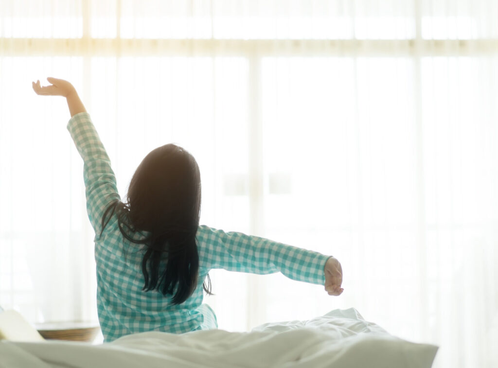 朝の光の中で伸びをする女性。自律神経が整い、心地よく目覚めるイメージ