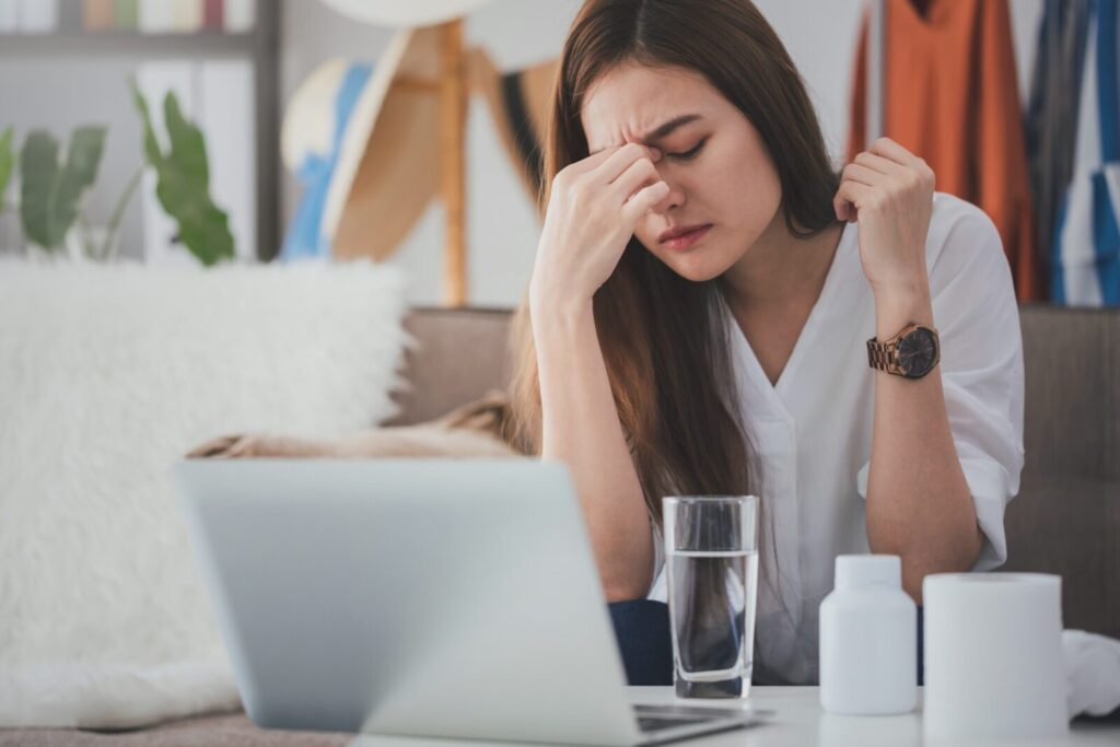 ノートパソコンの前で頭を抱える女性。めまいや頭痛に悩み、疲労感を感じている様子。